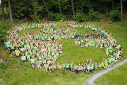 Viele naturbegeisterte Kinder mit grünene T-Shirts sitzen im Kreis und bilden gemeinsma das Logo des Nationalparks Sächsishce Schweiz ab.